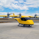 An air taxisitting at an airport runway under a blue sky. The Wisk Gen 6 is yellow with a black windshield and six propellers on top.